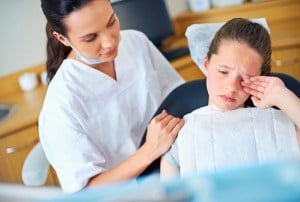 A young girl looking anxious and wiping her eye in a dental chair, with a comforting dental professional by her side, representing dental anxiety